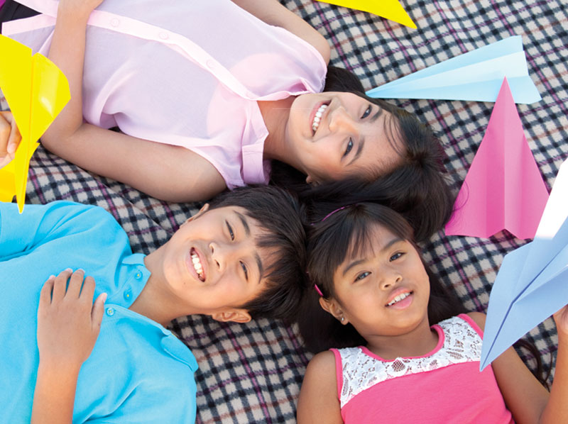 Two girls and a boy lay on a blanket and hold paper planes.